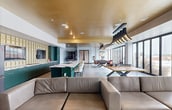 The Pool Lounge at Katz On Main spans 1,810 square feet on a single floor, offering a sophisticated and multifunctional amenity space designed for socializing and recreation. Upon entry, guests are welcomed by a vibrant foyer accented with bold artwork, a fuchsia accent chair, and large windows that flood the space with natural light. The lounge features large-format tile flooring throughout and a striking gold-painted tray ceiling that adds warmth and elegance to the interior. A centerpiece billiards table, finished in sleek black, is dramatically lit by a sculptural pendant light fixture and set atop an abstract area rug, anchored by walls of floor-to-ceiling windows that offer expansive outdoor views. Adjacent to the game area, a generous lounge zone provides plush neutral sectional seating, low coffee tables, and wall-mounted flat-screen televisions, complemented by bold framed artwork. The kitchen and bar area is outfitted with deep green cabinetry, a marble-topped island with bar seating, a full-size stainless steel refrigerator, and decorative tile backsplash detailing. A built-in bookshelf adds a curated residential touch. The space seamlessly balances entertainment, comfort, and contemporary design throughout.