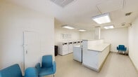 This single-floor shared laundry facility in Baton Rouge, Louisiana is designed for functional, communal use. The space is finished with neutral white walls and durable tile flooring, creating a clean and utilitarian environment throughout. Bright fluorescent panel lighting illuminates the interior evenly, ensuring excellent visibility across all areas of the room.

The laundry area is well-equipped with multiple front-loading and top-loading washing machines and dryers arranged along the walls, providing ample capacity for users. The machines feature digital control panels and appear to operate on a card or coin-based payment system, with posted instructional guides displayed on the wall for user reference. A continuous white folding counter runs along one side of the room, offering generous workspace for sorting and folding laundry.

A small seating area near the entrance provides a comfortable waiting space, furnished with blue upholstered chairs. A trash receptacle is conveniently positioned near the machines. The room is accessed through a standard interior door, with a second door visible at the far end. The layout is open and efficiently organized, maximizing usable floor space while maintaining clear circulation pathways throughout the facility.