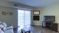This single-floor living space offers a well-appointed layout centered around a clean, functional living area. The room features hardwood-style flooring throughout, lending warmth and continuity to the space. A corner fireplace serves as a focal point, framed in white with a decorative mirror mounted above, adding depth and visual interest to the wall. Natural light is a defining characteristic of the space, streaming in generously through a large sliding glass door fitted with vertical blinds, which also provides access to an outdoor area with views of the surrounding neighborhood. The neutral, light-toned walls contribute to an open and airy atmosphere. A glass-top coffee table anchors the seating area, complementing the space's understated, contemporary aesthetic. A television stand with a wine rack detail is positioned near the fireplace wall, offering both function and style. Ceiling vents indicate a central HVAC system for year-round comfort. Overall, the space balances practicality with a welcoming ambiance, making it a comfortable and versatile living environment.