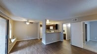 This single-floor residence offers a well-designed open-concept layout that maximizes the sense of space and functionality. The living area features rich, dark hardwood-style flooring that runs throughout the main space, complemented by neutral gray walls and crisp white baseboards that create a clean, modern aesthetic. A ceiling fan is centrally positioned in the living area, providing both comfort and a subtle design accent.

The kitchen is defined by a peninsula counter with a stone-look surface, offering both additional workspace and a natural divider between the kitchen and living areas. Dark espresso cabinetry lines the upper and lower kitchen walls, providing ample storage, while white appliances — including a refrigerator and built-in microwave — add a classic contrast. A textured popcorn ceiling adds a traditional touch throughout the space.

Adjacent to the main living area, a hallway leads to what appears to be a bathroom, featuring a window that brings in natural light, and additional cabinetry. The open sightlines between the kitchen, living, and adjoining spaces make this residence feel connected and functional, ideal for both everyday living and casual entertaining.