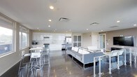 The Sky Lounge is a contemporary, open-plan communal space designed for socializing and relaxation. The layout flows seamlessly across a single level, with rich dark hardwood flooring grounding the bright, modern aesthetic throughout. Recessed lighting illuminates the space from above, complementing the clean white ceilings and neutral-toned walls.

The fully equipped kitchen is a standout feature, showcasing sleek, high-gloss white cabinetry, a stylish mosaic tile backsplash, and stainless steel appliances including a refrigerator, stove, and microwave. The kitchen opens directly to a casual dining area furnished with light wood-topped high tables and modern bar stools, creating an inviting spot for informal meals or conversation.

Adjacent to the kitchen, the lounge area is furnished with contemporary gray sectional sofas arranged around a central coffee table, offering a comfortable and stylish setting for gatherings. Frosted glass panel doors and interior glazing allow light to filter through while maintaining a sense of openness. The overall ambiance is clean, bright, and modern — a well-appointed communal retreat that balances function with a sophisticated, welcoming atmosphere.