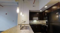 This single-floor space blends modern finishes with industrial-inspired design to create a distinctive and functional living environment. The open-concept kitchen serves as a central focal point, showcasing rich dark espresso cabinetry that extends to impressive heights, complemented by light granite countertops that provide a striking contrast. A peninsula with a stainless steel sink and granite surface extends into the living area, offering casual seating and a natural division between the kitchen and the rest of the space. Pendant lighting in cylindrical glass fixtures hangs dramatically from the lofty ceiling, casting warm, focused light over the peninsula and enhancing the space's modern aesthetic. The kitchen is fully equipped with black appliances, including a refrigerator, range, and built-in microwave. Soaring ceilings with exposed piping and ductwork underscore the loft-inspired character of the space, while recessed lighting provides additional illumination throughout. Polished concrete floors add to the urban, contemporary feel. A louvered natural wood interior door adds a warm, textural element that softens the otherwise bold palette of dark cabinetry, white walls, and gray trim throughout the space.
