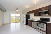 This single-floor unit offers a practical and cohesive layout designed for comfortable everyday living. The kitchen is a standout feature, outfitted with a full suite of stainless steel appliances including a refrigerator, range, microwave, and dishwasher. Rich, dark-stained cabinetry with brushed metal hardware provides generous storage both above and below the light-colored countertops, while a double-basin sink adds functional workspace. Flush-mounted industrial-style light fixtures illuminate the kitchen area effectively.

The open floor plan connects the kitchen to the main living area, which benefits from a ceiling fan and smooth, neutral-toned flooring throughout. The bedroom is bright and airy, featuring a large wood-framed window that allows natural light to fill the room and offers views of the surrounding neighborhood's tree-lined streetscape. A ceiling fan with a light fixture provides additional comfort and ventilation.

The bathroom is accessed through light wood-framed doors and features a clean, straightforward design with a toilet, vanity, and built-in shelving. An in-unit washer is also present, adding everyday convenience. Neutral wall tones and consistent flooring throughout the unit create a unified, clean aesthetic.