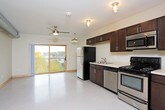 This single-floor unit offers a practical and cohesive layout designed for comfortable everyday living. The kitchen is a standout feature, outfitted with a full suite of stainless steel appliances including a refrigerator, range, microwave, and dishwasher. Rich, dark-stained cabinetry with brushed metal hardware provides generous storage both above and below the light-colored countertops, while a double-basin sink adds functional workspace. Flush-mounted industrial-style light fixtures illuminate the kitchen area effectively.

The open floor plan connects the kitchen to the main living area, which benefits from a ceiling fan and smooth, neutral-toned flooring throughout. The bedroom is bright and airy, featuring a large wood-framed window that allows natural light to fill the room and offers views of the surrounding neighborhood's tree-lined streetscape. A ceiling fan with a light fixture provides additional comfort and ventilation.

The bathroom is accessed through light wood-framed doors and features a clean, straightforward design with a toilet, vanity, and built-in shelving. An in-unit washer is also present, adding everyday convenience. Neutral wall tones and consistent flooring throughout the unit create a unified, clean aesthetic.