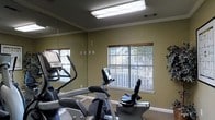 This fitness center is a dedicated, single-floor exercise space designed to support an active lifestyle. The room is outfitted with multiple cardio machines, including an elliptical trainer and a recumbent stationary bike, providing a range of workout options for users of varying fitness levels. A large floor-to-ceiling mirror spans one wall, creating a sense of expanded space while allowing users to monitor their form during exercise. The walls are finished in a warm olive-green tone, complemented by white crown molding that adds a refined touch to the utilitarian space. Overhead, a flush-mounted fluorescent light panel provides bright, even illumination throughout the room. Natural light filters in through two windows dressed with horizontal blinds, offering views of lush trees outside and creating an inviting, airy atmosphere. The carpeted flooring adds a layer of comfort underfoot, and a decorative plant near the window introduces a refreshing natural element. The overall layout is clean and functional, making efficient use of the available space while maintaining a welcoming environment conducive to regular exercise.