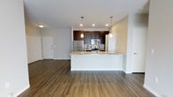 This single-floor residence showcases a thoughtfully designed open-concept layout that integrates the kitchen and living areas into one cohesive, flowing space. The kitchen is a standout feature, outfitted with rich dark espresso cabinetry that contrasts elegantly against light granite countertops. Stainless steel appliances, including a refrigerator, range, and microwave, add a modern, polished aesthetic. A white peninsula extends into the living space, creating a natural gathering point ideal for casual dining or entertaining.

Two pendant lights hang above the peninsula, providing focused task lighting that complements the recessed ceiling lights throughout. The wide-plank hardwood-style flooring runs continuously across the entire space, enhancing the sense of openness and visual cohesion. Neutral-toned walls in a soft gray-beige palette provide a versatile backdrop adaptable to a variety of interior styles.

The entry door is visible from the main living area, contributing to an intuitive and accessible floor plan. The overall ambiance is clean, contemporary, and well-lit, making the space feel both welcoming and functional.