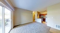 This single-floor space offers an open-concept design that seamlessly connects the living area to the kitchen. The living area is finished with wall-to-wall carpet in a neutral tone, complemented by beige walls that create a clean, understated backdrop. A wall-mounted climate control unit provides convenient temperature regulation.

The kitchen is situated adjacent to the living space and is defined by warm, dark wood cabinetry that extends to the upper walls, paired with light-colored countertops and a peninsula that serves as a natural divider between the two areas. The kitchen is equipped with stainless steel appliances, including a range with oven, an over-the-range microwave, and a full-sized refrigerator. Track lighting installed on the kitchen ceiling provides focused, adjustable illumination. The kitchen flooring transitions to a hard surface, contrasting with the carpeted living area.

A hallway visible from the living space leads to additional rooms within the unit, suggesting a practical and efficient floor plan. Overall, the space presents a functional and cohesive layout suited for everyday living.