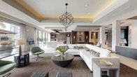 This elegantly designed single-floor space showcases a seamless blend of contemporary architecture and refined interior design. The open-concept layout flows effortlessly between distinct functional zones, creating a cohesive and inviting environment throughout.

The central lounge area is anchored by a generous sectional sofa in crisp white upholstery, complemented by a sculptural geometric chandelier suspended from a coffered ceiling with warm cove lighting. Large floor-to-ceiling windows and sliding glass doors flood the space with natural light and provide views of lush outdoor greenery.

A fully equipped demonstration kitchen features an expansive marble island with bar seating, sleek black pendant lighting, stainless steel appliances, and a textured tile backsplash — all framed by additional floor-to-ceiling windows that continue the indoor-outdoor connection.

The media and entertainment area is highlighted by a sophisticated dark wood feature wall housing a large flat-screen display framed in stone tile, flanked by elegant wall sconces and built-in display shelving adorned with organic décor elements.

A flexible lounge corridor incorporates a dramatic chain-curtain room divider, high-top tables, and mixed seating, ideal for casual gatherings. A marble-topped reception counter with sculptural accents anchors the entry zone, while curated artwork and neutral large-format tile flooring unify the space with a polished, contemporary ambiance.