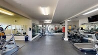 Located at 2040 SW Vermont St in Portland, Oregon, this fitness center offers a single-floor exercise space designed to support a comprehensive workout experience. The layout is open and functional, with durable rubber flooring throughout that provides both safety and comfort during exercise.

The facility is equipped with a wide variety of cardio machines, including treadmills, ellipticals, and stair climbers, making it suitable for sustained aerobic training. Strength and resistance training is well-represented with multi-station cable machines, weight benches, free weights, and a heavy bag for boxing workouts. Additional functional fitness equipment such as stability balls and wall-mounted accessories rounds out the offering.

Large mirrors line the walls, visually expanding the space and allowing users to monitor their form during exercise. The interior is illuminated by overhead fluorescent lighting, ensuring a bright and practical environment. Structural columns divide the floor plan into distinct zones without impeding movement, contributing to an organized flow throughout the space.

Natural light filters in through windows near the entrance, adding a welcoming quality to the otherwise utilitarian setting. A wall-mounted television and ceiling fans enhance comfort during use. Overall, this fitness center is thoughtfully arranged to provide residents with convenient access to a broad range of exercise options in a clean, well-maintained environment.