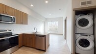 This thoughtfully designed unit at 420 W Stuart Rd in Bellingham, Washington, offers a clean, modern aesthetic with warm wood tones and light, neutral finishes throughout. The open-concept kitchen is a standout feature, equipped with stainless steel appliances including a refrigerator, electric range, and over-the-range microwave. Warm wood cabinetry extends to the ceiling, providing generous storage, while the white quartz peninsula and countertops offer ample workspace and casual dining potential. Light wood-style flooring runs through the main living areas, contributing to a bright and airy atmosphere. A large window in the living area frames natural views and draws in abundant natural light. The bedroom is carpeted for comfort and features a sizable window overlooking a wooded landscape, creating a peaceful, nature-connected retreat. The bathroom is finished with a full soaking tub and shower combination, a wood-cabinet vanity with a white countertop sink, and a large mirror. A stacked washer and dryer unit is conveniently included within the space. Recessed lighting throughout adds a polished, contemporary touch to every room.