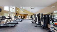 This single-floor fitness center offers a comprehensive workout environment designed to accommodate a wide range of exercise needs. The main workout area is defined by striking vaulted ceilings that rise dramatically overhead, creating an open and airy atmosphere enhanced by multiple ceiling fans that ensure comfortable air circulation throughout. Warm golden-yellow walls and plush carpeted flooring add a welcoming feel to the space.

The cardio zone features a row of treadmills and an elliptical trainer, while the strength training area is outfitted with multiple cable and weight machines, a dedicated free weights rack stocked with dumbbells, kettlebells, and barbells, and adjustable benches for varied exercises. Large windows line the perimeter, flooding the space with natural light and offering views of the surrounding greenery and outdoor pool area.

A separate, more intimate exercise room provides space for stretching and flexibility work, complete with stability balls and additional equipment. A wood-paneled sauna is also available for post-workout recovery. Full-length mirrors along the walls further enhance the sense of space while supporting proper form during training. The facility is thoughtfully laid out, balancing open floor space with a thorough selection of equipment for a well-rounded fitness experience.