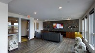 This contemporary single-floor lounge space is thoughtfully designed to offer both comfort and sophistication. The centerpiece of the room is a striking feature wall clad in dark mosaic tile, which frames a large wall-mounted flat-screen television and serves as a backdrop for an elegant built-in display area showcasing wine bottles and decorative items. Warm wood-toned lower cabinetry with a built-in bar sink runs along the base of the feature wall, providing both functional storage and an entertainment-ready setup. The seating area is anchored by a sleek gray leather sofa and an accent chair, arranged to face the screen — ideal for gatherings or relaxed viewing. Recessed and track lighting throughout the ceiling provides layered illumination, enhancing the room's modern ambiance. The flooring consists of large-format wood-look porcelain tile, lending a clean and upscale aesthetic that ties the space together. Framed artwork adorns the white walls, adding a curated touch to the interior. A glass-paneled door provides access to an adjacent area, contributing to the open and connected feel of this well-appointed lounge.