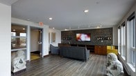 This contemporary single-floor lounge space is thoughtfully designed to offer both comfort and sophistication. The centerpiece of the room is a striking feature wall clad in dark mosaic tile, which frames a large wall-mounted flat-screen television and serves as a backdrop for an elegant built-in display area showcasing wine bottles and decorative items. Warm wood-toned lower cabinetry with a built-in bar sink runs along the base of the feature wall, providing both functional storage and an entertainment-ready setup. The seating area is anchored by a sleek gray leather sofa and an accent chair, arranged to face the screen — ideal for gatherings or relaxed viewing. Recessed and track lighting throughout the ceiling provides layered illumination, enhancing the room's modern ambiance. The flooring consists of large-format wood-look porcelain tile, lending a clean and upscale aesthetic that ties the space together. Framed artwork adorns the white walls, adding a curated touch to the interior. A glass-paneled door provides access to an adjacent area, contributing to the open and connected feel of this well-appointed lounge.