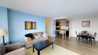 This 1-bedroom, 1-bathroom unit is designed with an open-concept layout that seamlessly connects the living area and kitchen, creating a functional and inviting space. The living room features warm hardwood flooring, a light gray sofa accented with colorful throw pillows, and a bold sky-blue accent wall that adds a vibrant focal point to the room. A framed wall art piece and a sleek black coffee table complete the living space. The kitchen is equipped with dark wood cabinetry, black appliances, and a granite-topped breakfast bar on a rolling island, offering flexible dining and prep options. Track lighting above the kitchen provides focused illumination, while recessed lighting throughout ensures the space remains bright and well-lit. White walls and ceilings contrast cleanly with the darker cabinetry and flooring, contributing to a crisp, modern aesthetic. The overall layout is compact yet efficiently organized, making excellent use of the available space to accommodate everyday living with ease.