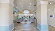 The Pacific Islands clubhouse is a single-story community facility designed with a bright, coastal aesthetic that carries consistently throughout the space. The grand entrance welcomes visitors through glass double doors topped by a striking arched window, flooding the foyer with natural light and setting an open, airy tone. Wood-look flooring extends throughout, complementing the soft blue and neutral beige color palette that defines the interior.

The main lounge area is the centerpiece of the clubhouse, featuring soaring vaulted ceilings with recessed lighting and a dramatic arched architectural detail overhead. Comfortable gray sofas accented with teal and gold throw pillows are arranged around a glass-top coffee table, creating a relaxed seating area. A standout feature wall clad in mosaic tile houses both a working fireplace and a wall-mounted flat-screen television, adding warmth and character to the space.

Adjacent seating areas offer additional comfort, decorated with coastal-inspired framed artwork and wicker accent chairs. A pass-through kitchenette provides practical amenities, equipped with stainless steel appliances, rich wood cabinetry, and ample counter space. Large windows throughout the clubhouse invite natural light and views of the surrounding landscape. A fitness center, visible through glass partitions, rounds out the facility's offerings, making this clubhouse a well-appointed and functional community gathering space.
