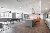 The community room at 2930 Rockaway Ave in Oceanside, NY is a thoughtfully designed, open-plan gathering space that seamlessly blends recreation, relaxation, and socializing. Wood-look tile flooring runs throughout, lending warmth and continuity across each distinct zone. A dropped tray ceiling with recessed lighting and brushed brass pendant fixtures adds architectural interest while ensuring the space is well-lit and inviting.

The lounge area is anchored by a cluster of rich caramel leather armchairs arranged around a central coffee table, with a large wall-mounted flat-screen TV set against a striking textured accent wall featuring an abstract, multicolored tile pattern. Adjacent to the lounge, a full-size billiards table rests on a patterned area rug, offering a dedicated recreation zone with ample room to play.

The kitchenette is well-equipped with light blue-gray cabinetry, a mosaic tile backsplash, open wood shelving, a stainless steel refrigerator, microwave, and sink — ideal for hosting events or casual gatherings. A large central island with a clean white countertop provides additional prep and serving space.

A casual dining area rounds out the room, featuring round white tables and modern white and gray chairs beneath industrial-style pendant lights, with natural light streaming in through windows. The overall ambiance is modern, clean, and community-focused.