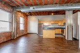 Located in the heart of Kansas City, MO, this urban loft showcases a compelling blend of industrial heritage and contemporary living. The open-concept main living area is defined by striking exposed brick walls, soaring ceilings with original tile and beam construction, and prominent ductwork that highlights the building's industrial roots. Rich hardwood flooring extends throughout the main living spaces, while track lighting and a ceiling fan add both function and style.

The kitchen is well-appointed with light maple cabinetry, dark granite-style countertops, stainless steel appliances including a refrigerator, range, and microwave, and a central island that provides additional workspace and storage. Tile flooring grounds the kitchen area, clearly delineating it from the living spaces.

The carpeted areas offer a softer, quieter retreat within the loft, with multiple windows along the brick walls allowing natural light to filter throughout. The bathroom features a full tub and shower combo, a wood-toned vanity with ample counter space, and tile flooring. A dedicated laundry area includes a full-size washer and dryer with overhead cabinetry and built-in shelving for added storage convenience. This space offers an authentic urban loft experience in a vibrant Kansas City location.