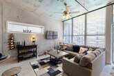 This single-floor urban loft showcases a bold industrial-chic design defined by raw exposed concrete ceilings and walls throughout. The open-concept layout seamlessly connects the living, dining, and kitchen areas, creating a cohesive and spacious feel enhanced by polished concrete floors that run throughout the unit.

The living area is a standout, featuring soaring ceilings with a ceiling fan, large floor-to-ceiling windows dressed with horizontal blinds that flood the space with natural light. A wall-mounted flat-screen TV and a bar cart complete the relaxed yet stylish atmosphere.

The kitchen is equipped with warm-toned wood cabinetry, dark countertops, a tile backsplash, and black appliances including a range and refrigerator. Track lighting and a bold navy blue accent wall add personality and definition to this functional galley-style kitchen.

A dedicated dining nook with a round table and industrial-style stools sits adjacent to the kitchen, providing a comfortable space for casual dining.

The entryway corridor features dramatic exposed concrete walls that set the tone for the loft's aesthetic from the moment you step inside. The bathroom offers a clean and functional design with wood vanity cabinetry, dark countertops, a framed mirror, and a shower-tub combination with a decorative curtain and a high window for natural light.