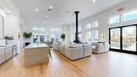 Located at 72 Eagle Rock Ave in East Hanover, NJ, this single-floor community amenity space offers a thoughtfully designed, open-concept layout that maximizes both functionality and aesthetic appeal. The space is characterized by soaring double-height ceilings that amplify its generous square footage, creating an airy and expansive atmosphere throughout.

The living area serves as the centerpiece, anchored by a dramatic black suspended pendant fireplace that descends from the ceiling, adding a bold architectural focal point. Light hardwood flooring flows seamlessly throughout the entire space, complementing the crisp white walls and recessed lighting overhead. Large windows positioned at both standard and clerestory heights flood the interior with natural light, while black-framed double doors provide direct access to an outdoor terrace.

A sleek, light wood island with a white quartz countertop defines the lounge and gathering area, which features generous sectional seating arrangements ideal for socializing. A dedicated game and recreation area includes a foosball table and wall-mounted TV, accompanied by modern accent seating. A wet bar with cabinetry and contemporary artwork adds a refined finishing touch. The overall ambiance is modern and minimalist, with a clean, neutral palette that conveys sophistication and comfort in equal measure.