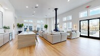 Located at 72 Eagle Rock Ave in East Hanover, NJ, this single-floor community amenity space offers a thoughtfully designed, open-concept layout that maximizes both functionality and aesthetic appeal. The space is characterized by soaring double-height ceilings that amplify its generous square footage, creating an airy and expansive atmosphere throughout.

The living area serves as the centerpiece, anchored by a dramatic black suspended pendant fireplace that descends from the ceiling, adding a bold architectural focal point. Light hardwood flooring flows seamlessly throughout the entire space, complementing the crisp white walls and recessed lighting overhead. Large windows positioned at both standard and clerestory heights flood the interior with natural light, while black-framed double doors provide direct access to an outdoor terrace.

A sleek, light wood island with a white quartz countertop defines the lounge and gathering area, which features generous sectional seating arrangements ideal for socializing. A dedicated game and recreation area includes a foosball table and wall-mounted TV, accompanied by modern accent seating. A wet bar with cabinetry and contemporary artwork adds a refined finishing touch. The overall ambiance is modern and minimalist, with a clean, neutral palette that conveys sophistication and comfort in equal measure.