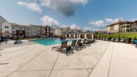 Located at 1062 W Oakland Ave in Johnson City, Tennessee, this outdoor amenity area is centered around a generously sized resort-style swimming pool that serves as the focal point of the community's recreational space. The pool features a distinctive shallow wading shelf lined with in-water chaise loungers, allowing residents to relax partially submerged, as well as a decorative water fountain that adds a dynamic visual element to the space.

Surrounding the pool is a spacious concrete deck thoughtfully appointed with an abundance of lounge chairs, many shaded by blue market umbrellas, offering a variety of sun and shade options throughout the day. A wood-framed pergola anchors one corner of the deck, providing a shaded gathering spot with additional seating for socializing or unwinding. A private cabana with curtained sides offers a more secluded retreat along the pool's edge.

Outdoor dining areas with chairs and tables are positioned near the perimeter, complemented by lush landscaping and a black iron fence that borders the space. The pool area enjoys open sky views and overlooks the broader residential community, with multi-story buildings and rolling greenery visible in the background. The overall design is polished and resort-inspired, delivering a welcoming and well-equipped outdoor living environment.