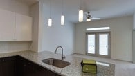 This single-floor residence offers a thoughtfully designed open-concept layout that connects the kitchen and living areas in a bright, cohesive space. The kitchen is appointed with granite countertops, dark cabinetry, and a stainless steel sink with a gooseneck faucet. Three sleek cylindrical pendant lights hang above the kitchen island, providing focused task lighting while adding a contemporary aesthetic touch. The soaring ceilings enhance the sense of openness and volume throughout the main living area, which is further complemented by a ceiling fan that promotes comfortable airflow. Neutral, light-toned walls create a clean and versatile backdrop throughout the space. The living area transitions smoothly toward a set of glass-panel double doors that flood the interior with natural light and provide direct access to the outdoors, offering a seamless indoor-outdoor connection. The overall ambiance is modern and airy, with a neutral palette and quality finishes that lend a polished yet inviting atmosphere. The combination of functional design and natural light makes this a well-appointed, comfortable living space.