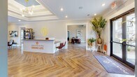 The Pointe Grand leasing office on Pointe Grand Drive in Augusta, Georgia, offers a well-appointed, single-story space designed to make a strong first impression. Guests are welcomed at a modern reception desk emblazoned with "Welcome to the Grand Life," set against warm herringbone-patterned hardwood flooring that extends throughout the entire space. Crown molding and recessed lighting add a polished, refined quality to the entry area, which is further accented by lush greenery and curated décor.

Moving deeper into the space, the main lounge area commands attention with a striking burnt-orange shiplap accent wall, exposed dark wood beam ceilings, and vivid teal-blue painted walls. Comfortable leather seating, a decorative media console, and built-in bookshelves create an inviting, residential atmosphere. Large windows allow natural light to filter in, offering views of the surrounding community.

Adjacent areas include a casual dining nook with a round wooden table and leather chairs, as well as a private office space visible through glass-paneled doors. Bold artwork throughout reinforces the vibrant, design-forward aesthetic. The overall ambiance is energetic yet welcoming, reflecting a thoughtfully curated environment.