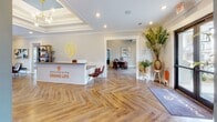 The Pointe Grand leasing office on Pointe Grand Drive in Augusta, Georgia, offers a well-appointed, single-story space designed to make a strong first impression. Guests are welcomed at a modern reception desk emblazoned with "Welcome to the Grand Life," set against warm herringbone-patterned hardwood flooring that extends throughout the entire space. Crown molding and recessed lighting add a polished, refined quality to the entry area, which is further accented by lush greenery and curated décor.

Moving deeper into the space, the main lounge area commands attention with a striking burnt-orange shiplap accent wall, exposed dark wood beam ceilings, and vivid teal-blue painted walls. Comfortable leather seating, a decorative media console, and built-in bookshelves create an inviting, residential atmosphere. Large windows allow natural light to filter in, offering views of the surrounding community.

Adjacent areas include a casual dining nook with a round wooden table and leather chairs, as well as a private office space visible through glass-paneled doors. Bold artwork throughout reinforces the vibrant, design-forward aesthetic. The overall ambiance is energetic yet welcoming, reflecting a thoughtfully curated environment.