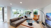 Located on the 3rd floor of a contemporary Vancouver residential building on Davie Street, this amenity room offers a thoughtfully designed communal space that blends modern interiors with generous outdoor living areas. The interior is finished with light hardwood flooring throughout, recessed lighting, and clean white walls that create a bright, open atmosphere. The living area is furnished with a modular sectional sofa, a classic leather egg chair, an arc floor lamp, and a round coffee table — all centered around a large flat-screen television, making it an ideal space for relaxed gatherings. Floor-to-ceiling windows flood the lounge with natural light and frame lush greenery from the surrounding terraces. The kitchen features sleek, handleless matte dark cabinetry contrasted with white upper cabinets and a marble-effect backsplash. A substantial dark-topped island with bar seating serves as a central gathering point, complemented by built-in appliances and ample counter space. Outside, multiple terrace areas extend the amenity offering, including a landscaped deck with decorative rock features and a built-in BBQ station, a spacious turf lawn with glass-railed perimeter views of the Vancouver skyline and surrounding greenery, and a communal dining terrace with long wooden tables and cube seating beneath string lights.