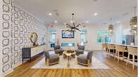 Located at 185 Rosewood Loop in Burlington, NC, this thoughtfully designed single-floor space combines functionality with contemporary style. The layout encompasses multiple living areas, a well-appointed kitchen, a dedicated office, and a connecting hallway, all unified by warm hardwood-style flooring and a bright, welcoming palette.

The main living area serves as the centerpiece of the space, featuring a striking bold-patterned geometric accent wall, a sleek linear fireplace framed in white shiplap, and a wall-mounted flat-screen TV. A sculptural multi-arm chandelier anchors the seating arrangement of modern upholstered chairs gathered around a round marble-top coffee table. Additional living areas provide flexible seating and gathering options, with natural light filtering in through multiple windows.

The kitchen is outfitted with white shaker cabinetry accented by gold hardware, granite countertops, a dark subway tile backsplash, stainless steel appliances, and a central island with bar seating. Pendant lighting with woven gold fixtures adds warmth and elegance.

The private office is a quiet, carpeted retreat with a built-in desk, two workstations, and a large window overlooking the outdoor surroundings. The hallway entry area features tasteful décor and a glass-panel door that welcomes natural light into the space.
