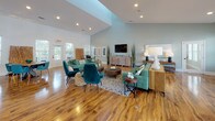 Located at 5418 Sirius Dr in Wilmington, NC, this single-floor community clubhouse is designed with an open-concept layout that seamlessly connects its various functional areas. Rich wood-look flooring flows throughout the entire space, complementing soft, muted wall tones of grey and sage green that lend a calm, coastal ambiance.

The kitchen is a standout feature, outfitted with white shaker cabinetry, granite countertops, stainless steel appliances including a refrigerator and range with a wall-mounted vent hood, and a generous peninsula with seating for multiple bar-height chairs. Stylish pendant lights hang above the counter, while a built-in beverage station with additional counter space and under-counter appliances serves as a secondary prep and refreshment area.

The lounge area is expansive, with soaring vaulted ceilings and abundant natural light streaming through multiple large windows. Comfortable seating arrangements in teal and neutral tones, along with a wall-mounted television and coastal artwork, create a welcoming gathering environment. A round dining table with teal chairs and decorative driftwood accents adds a natural, beachy character to the dining zone.

A dedicated office area provides workstations, natural light, and private access to a smaller adjoining room. Throughout, recessed lighting and large windows ensure the space remains bright and inviting.
