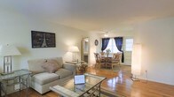 This single-floor space presents an open-plan layout that seamlessly connects the living and dining areas, creating a welcoming and functional environment. The living area is anchored by a plush, neutral-toned sofa paired with a glass-top coffee table, adding a touch of elegance while maintaining an airy feel. Decorative accents, including a Paris-themed wall art piece, lend the space a casual yet stylish character.

Warm hardwood floors run throughout, adding richness and continuity between the two spaces. A floor lamp provides soft ambient lighting in the living area, complementing the natural light that filters in through the dining room's curtained windows. The dining area is furnished with a round table and rattan-style chairs, evoking a relaxed, casual dining experience. A ceiling fan overhead ensures comfort year-round.

A mirrored accent near the transition between rooms enhances the sense of depth and light. The neutral wall tones throughout create a clean backdrop that allows the furnishings and décor to stand out. Overall, the space offers a comfortable and cohesive atmosphere suitable for everyday living and entertaining.