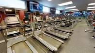 This fitness center offers a comprehensive workout environment designed to meet a wide range of exercise needs. The cardio zone is a standout feature, lined with rows of modern treadmills and elliptical machines, each equipped with individual display consoles for tracking performance metrics. Large flat-screen televisions are mounted along the walls, allowing members to stay entertained or informed during their workouts. Mirrored walls run the length of the cardio area, creating a sense of expansiveness while allowing exercisers to monitor their form. The flooring throughout is durable rubber, providing both safety and comfort underfoot. Natural light filters in through horizontal windows positioned along the upper portion of the walls, complementing the recessed and panel ceiling lighting to ensure the space is well-lit at all times. The bold accent walls in deep red add a dynamic, energizing aesthetic to the environment. Beyond the cardio section, additional equipment is visible in the background, suggesting a broader range of strength and conditioning options available throughout the facility. The overall ambiance is clean, organized, and purposefully designed to motivate and support an active fitness routine.