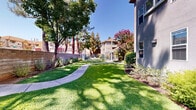 Located along El Camino Real in Santa Clara, California, this multi-building residential property offers a thoughtfully designed outdoor environment that balances comfort and functionality. The exterior features a stucco facade with a neutral gray finish, complemented by mature trees and carefully maintained shrub beds that frame the grounds with year-round greenery.

The expansive common areas are a defining feature of the property. A gently curving walkway winds through lush, manicured lawns shaded by towering pine and deciduous trees, creating a peaceful, park-like atmosphere. Colorful flowering shrubs, including blooming oleanders, add vibrant seasonal interest throughout the landscape.

Residents have access to a private spa enclosed by a secure wrought-iron fence, surrounded by mature trees that provide natural shade and privacy. Adjacent outdoor seating and lounge areas invite relaxation in the California sunshine. A dedicated picnic area with benches offers a welcoming space for outdoor gatherings.

The 3D aerial view of the property reveals two primary residential buildings set along a linear footprint, with generous green space and mature landscaping interspersed between them, connected by a network of paved pathways. The overall ambiance of the property is serene and well-appointed, reflecting the mild Silicon Valley climate and a commitment to comfortable outdoor living.
