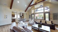 This single-story space makes a striking impression with its dramatic vaulted ceiling, defined by rich, dark-stained exposed wood beams that draw the eye upward. The open-concept layout creates a seamless connection between the main living area, a casual dining space, and the kitchen beyond, promoting an effortless flow throughout.

The living area is generously proportioned, accommodating multiple seating arrangements including dark leather sofas and neutral upholstered accent chairs centered around a wooden coffee table. Warm hardwood-style flooring runs throughout, grounding the space with a natural, cohesive aesthetic.

An abundance of natural light is a defining feature, delivered through a dramatic wall of tall windows and a transom arrangement that frames views of lush greenery and outdoor foliage. The windows also provide access to an outdoor area via a glass door.

The kitchen, visible in the background, features dark cabinetry, a pendant-lit island with bar seating, and a statement range hood, offering both functionality and style. Recessed lighting supplements natural light throughout, ensuring the warm, cream-toned walls remain well-illuminated at all times.