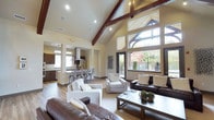 This single-story space makes a striking impression with its dramatic vaulted ceiling, defined by rich, dark-stained exposed wood beams that draw the eye upward. The open-concept layout creates a seamless connection between the main living area, a casual dining space, and the kitchen beyond, promoting an effortless flow throughout.

The living area is generously proportioned, accommodating multiple seating arrangements including dark leather sofas and neutral upholstered accent chairs centered around a wooden coffee table. Warm hardwood-style flooring runs throughout, grounding the space with a natural, cohesive aesthetic.

An abundance of natural light is a defining feature, delivered through a dramatic wall of tall windows and a transom arrangement that frames views of lush greenery and outdoor foliage. The windows also provide access to an outdoor area via a glass door.

The kitchen, visible in the background, features dark cabinetry, a pendant-lit island with bar seating, and a statement range hood, offering both functionality and style. Recessed lighting supplements natural light throughout, ensuring the warm, cream-toned walls remain well-illuminated at all times.