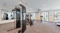 Located at 19939 Longenbaugh Rd in Houston, TX, this fitness center offers a comprehensive and well-appointed exercise facility designed to support a full range of workout routines. The single-floor space is generously sized, providing ample room for both equipment and free movement throughout.

The layout is thoughtfully organized into distinct workout zones. The strength training area features a Precor cable machine system, a multi-function power rack with bench press, adjustable benches, and a full dumbbell rack spanning a wide range of weights, complemented by kettlebells and medicine balls stored on dedicated racks. Large wall-mounted mirrors in this zone enhance the sense of space and allow for proper form monitoring.

The cardio section is equipped with multiple elliptical trainers, treadmills, and stationary cycling bikes — including a Peloton-style interactive cycle — all positioned along the windowed perimeter of the room to allow for natural light and outdoor views.

Underfoot, durable carpet tile flooring covers the entire space, providing comfort and traction. Recessed lighting and multiple ceiling fans ensure the room remains well-lit and well-ventilated. Large black-framed windows line multiple walls, flooding the interior with natural light and offering views of the surrounding outdoor greenery, creating an open and energizing workout environment.