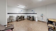Located at 4141 Bathurst Street in Toronto, Ontario, this shared laundry facility offers a practical and well-organized space for residents. The room is finished with durable terrazzo flooring throughout, white painted walls accented by a horizontal band of dark grey tile, and bright fluorescent overhead lighting that ensures excellent visibility.

The laundry area is equipped with multiple front-loading LG washers and dryers arranged along the walls, providing ample capacity for residents. Instructional signage for washer and dryer usage is clearly posted above the machines, and a wall-mounted clock keeps track of cycle times. A utility sink is available along one wall, and a payment station is conveniently positioned nearby.

A waiting area adjacent to the laundry machines offers comfortable seating, including armchairs and a small table, making it easy for residents to wait during their laundry cycles. A community bulletin board mounted on the wall displays notices and information relevant to building residents.

Two access doors provide entry and exit points to the space. The overall layout is efficient and straightforward, designed to accommodate multiple users simultaneously. The clean, minimal aesthetic and practical amenities make this a functional shared facility well-suited to the needs of a residential building.