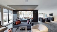 This elegantly designed single-floor space offers a refined and welcoming atmosphere through a thoughtful blend of contemporary aesthetics and functional layout. The interior is anchored by a bold dark architectural partition that defines distinct seating zones while maintaining an open, flowing sense of space throughout.

The main lounge area features plush, dark upholstered sofas accented with vibrant throw pillows in deep reds, blues, and purples, adding warmth and personality against the sophisticated dark and neutral palette. A patterned area rug grounds the primary seating arrangement, while a secondary lounge zone beyond the partition is bathed in natural light streaming through large windows, creating a bright and airy contrast.

Overhead, a pair of striking globe-shaped pendant lights serve as focal points, adding a sculptural element to the space. Recessed lighting complements the ambient illumination throughout. The secondary zone features additional lounge seating with an intimate, sunlit quality ideal for casual gatherings or relaxed conversation.

To one side, a row of individual armchairs and side tables creates a quieter, more private seating arrangement. The overall design balances drama and comfort, with dark wall paneling, layered textures, and carefully curated furnishings coming together to produce a polished, upscale environment suited for socializing and relaxation.