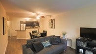 This single-floor residence offers an open-concept layout that brings together the main living areas in a cohesive and practical design. The living room serves as the heart of the space, featuring rich hardwood flooring, neutral-toned walls, and a ceiling fan that enhances comfort and airflow. A generously sized sectional sofa anchors the room, complemented by tasteful wall art that adds a decorative touch to the warm, inviting ambiance.

Flowing naturally from the living room, the dining area accommodates a full dining table and chairs, making it well-suited for both everyday meals and entertaining. The open sightlines between the dining and living spaces create a sense of continuity and ease of movement throughout.

The kitchen is positioned just beyond the dining area and is outfitted with dark cabinetry, stainless steel appliances — including a refrigerator and microwave — and a compact countertop workspace. The layout is efficient and functional, designed to support a variety of cooking needs.

Warm, ambient lighting throughout the main living areas contributes to the cozy and welcoming atmosphere of this space. The combination of hardwood flooring, contemporary furnishings, and a well-integrated open floor plan makes this a comfortable and stylish single-floor home.
