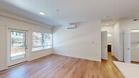 This single-floor unit offers a thoughtfully designed layout encompassing a combined kitchen and living area, one bedroom, one bathroom, and a hallway. The open-concept living area is characterized by light-toned hardwood-style flooring that runs throughout the unit, crisp white walls, and recessed lighting. A wall-mounted mini-split AC unit provides efficient climate control, while a glass-paneled door and an adjacent window allow natural light to fill the space and provide direct outdoor access. The kitchen is well-equipped with dark wood cabinetry, stainless steel appliances including a refrigerator, range, microwave, and dishwasher, a white subway tile backsplash, light quartz countertops, and a central island with pendant lighting overhead. The bedroom features its own window with outdoor views, a ceiling fan with light fixture, and continues the same hardwood-style flooring. The bathroom is finished with a dark wood vanity, light countertop, a large mirror, a tub-shower combination, and hardwood-style flooring consistent with the rest of the unit.