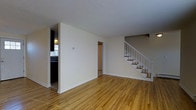 This two-floor space is defined by warm-toned hardwood flooring that extends throughout the main level, creating a cohesive and inviting atmosphere. The main living area is open and generously sized, with neutral cream-colored walls and a textured ceiling that together contribute to a clean, understated aesthetic. Natural light filters in from the adjacent kitchen area, which is partially visible through a half-wall partition to the left. The kitchen features dark cabinetry and provides a functional contrast to the bright, airy living space.

A centrally positioned staircase with white-painted railings and wooden treads leads to the second floor, serving as a notable architectural feature within the layout. The staircase placement creates a natural division between the living area and a hallway that leads further into the home. A doorway visible in the background hints at additional rooms beyond the main space. The overall design is practical and straightforward, with consistent finishes throughout. The combination of light walls, hardwood floors, and white trim gives the space a timeless, move-in-ready quality suitable for a variety of uses.