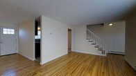 This two-floor space is defined by warm-toned hardwood flooring that extends throughout the main level, creating a cohesive and inviting atmosphere. The main living area is open and generously sized, with neutral cream-colored walls and a textured ceiling that together contribute to a clean, understated aesthetic. Natural light filters in from the adjacent kitchen area, which is partially visible through a half-wall partition to the left. The kitchen features dark cabinetry and provides a functional contrast to the bright, airy living space.

A centrally positioned staircase with white-painted railings and wooden treads leads to the second floor, serving as a notable architectural feature within the layout. The staircase placement creates a natural division between the living area and a hallway that leads further into the home. A doorway visible in the background hints at additional rooms beyond the main space. The overall design is practical and straightforward, with consistent finishes throughout. The combination of light walls, hardwood floors, and white trim gives the space a timeless, move-in-ready quality suitable for a variety of uses.