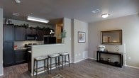 This single-floor residence showcases an open-concept design that seamlessly connects the kitchen and living areas, creating a cohesive and functional living space. The kitchen is equipped with dark espresso cabinetry, black appliances including a refrigerator and microwave, and granite-style countertops. A raised breakfast bar with seating for three cushioned barstools serves as a natural divider between the kitchen and the adjoining living space, making it ideal for casual dining or entertaining.

The flooring throughout is a warm-toned wood-look material that adds continuity and a modern touch across the open layout. Neutral beige walls provide a versatile backdrop, complemented by white baseboards and trim. A console table along one wall offers additional surface space, and framed wall art adds decorative character to the room. Recessed and flush-mount ceiling lighting ensures the space is well-lit throughout. Ventilation is provided through floor-level vents, and a thermostat is conveniently positioned on the wall. The overall ambiance is clean, contemporary, and well-organized, making efficient use of the available space.
