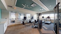 This fitness center is thoughtfully designed to support a comprehensive workout experience within a single, open-plan space. The interior is defined by a distinctive tray ceiling finished in teal, complemented by recessed lighting, ceiling fans, and crisp white trim that together create a polished, modern aesthetic. Patterned carpet tiles in a herringbone-inspired layout cover the floor throughout, adding visual interest while providing a durable, functional surface.

The cardio zone is well-appointed with multiple treadmills, elliptical trainers, and stationary bikes, each equipped with individual monitor displays. Large windows line the walls, flooding the space with natural light and offering views of the surrounding outdoor environment. Full-length mirrors along one wall visually expand the room and include a wall-mounted flat-screen television.

The strength training area includes a multi-functional cable machine, a complete dumbbell rack ranging across various weights, and a weight bench, providing versatile options for resistance training. Near the entrance, a refreshment station features a water fountain, built-in shelving, and cabinetry accented by a teal tile backsplash. Abstract wall art adds a touch of color and personality to the space. Glass entry doors allow sightlines into the fitness area from outside, reinforcing the open and welcoming atmosphere.