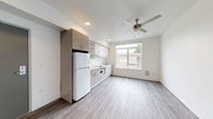 This thoughtfully designed single-floor unit offers a clean, contemporary aesthetic throughout. The open-concept main area seamlessly integrates the kitchen and living space, finished with light gray wood-look luxury vinyl plank flooring that runs throughout the entire unit. The kitchen is efficiently laid out along one wall, featuring two-tone cabinetry in white and warm taupe, a smooth cooktop, built-in microwave, full-size refrigerator, and a stainless steel sink — all complemented by white countertops and a large window that invites natural light. A ceiling fan overhead adds both comfort and style to the main living area, which offers generous open floor space ideal for flexible furniture arrangements. The bedroom is a well-proportioned, quiet retreat with its own ceiling fan, a window with an outdoor view, and ample wall space. An additional room provides versatile space suitable for a home office or storage. The bathroom is finished with the same durable vinyl flooring and includes a full vanity, toilet, and a shower-tub combination with chrome fixtures and grab bar. Recessed lighting and a neutral white palette throughout give the entire unit a bright, airy, and move-in-ready feel.