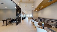Located in Miramar, FL, this single-floor clubhouse at 4970 SW 124th Ave offers a sophisticated and contemporary community gathering space. The main lounge corridor features soaring ceilings, large-format light tile flooring, and elegant gold pendant globe lights that draw the eye through the space toward floor-to-ceiling glass doors opening to outdoor views of lush tropical landscaping. A striking laser-cut black decorative partition adds an artistic focal point at the entrance.

Seating areas are thoughtfully varied: a tufted gray banquette with round dark tables and modern white chairs provides casual dining-style seating, while plush blush armchairs with gold legs offer a relaxed lounge setting. A dedicated lounge nook showcases dramatic vertical wood slat wall and ceiling detailing paired with deep charcoal sectional sofas and globe pendant lighting.

The game room features exposed wood beam ceilings, a pool table, casual seating, and direct outdoor access, creating a vibrant social hub. A coffee and refreshment bar with warm wood cabinetry and open shelving serves the space conveniently. The leasing or administrative area includes a reception desk with a textured accent wall and mounted display. A private conference room is finished with wood-paneled walls, a large dining table, upholstered seating, and a bold sculptural brass chandelier.