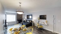 This single-floor space showcases a thoughtfully designed open-concept layout that seamlessly integrates the living and dining areas. The dining area is anchored by a glass-top table surrounded by striking yellow chairs, illuminated by a contemporary pendant light that draws the eye upward to the textured ceiling. The living area centers around a charcoal sectional sofa complemented by coordinating accent pillows and a tufted chaise, set atop a neutral area rug over durable hardwood-style flooring. A corner stone fireplace serves as a standout architectural feature, adding warmth and a natural texture that contrasts beautifully with the clean white walls. On the opposite side of the living area, a wall-mounted shelving and desk unit provides a functional workspace without compromising the open feel of the room. Bold yellow accent chairs and decorative accessories carry a consistent color story throughout the space. Natural light filters in through windows along the perimeter, contributing to the bright, airy atmosphere. The overall ambiance is modern and cohesive, balancing comfort with contemporary style.