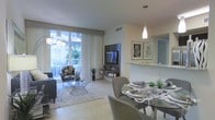 This single-floor residence features an open-concept layout that seamlessly integrates the living and dining areas into one cohesive space. The living area is anchored by a wall-mounted flat-screen TV on a sleek media console, complemented by contemporary furnishings including a plush sofa and accent chair arranged atop a soft area rug. Large sliding glass doors flood the room with abundant natural light while providing direct access to a private balcony, where views of tropical greenery create a serene outdoor backdrop. The dining area is positioned adjacent to the living space and features a glass-top round table surrounded by upholstered chairs, elegantly set and lit by a modern pendant light overhead. A half-wall with a stone ledge subtly separates the dining space from what appears to be a kitchen area beyond. The flooring throughout consists of neutral-toned large-format tiles that enhance the sense of openness. The overall color palette is light and airy, with crisp whites, warm grays, and natural accents contributing to a clean, contemporary ambiance throughout the space.