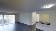 This single-floor space features an open-concept layout that seamlessly connects the living area and kitchen. The flooring throughout is a rich, dark hardwood-style material that contrasts cleanly with the bright white walls and neutral-toned ceiling, creating a modern and cohesive aesthetic. The living area is generously sized, offering a blank canvas with smooth, white walls and subtle baseboard trim. A sliding glass door along one wall floods the space with natural light while providing direct access to an outdoor patio or balcony area, enhancing the sense of openness. Adjacent to the living space, a kitchen counter with a dark laminate surface is partially visible, suggesting a functional kitchen area just beyond. A mirror or pass-through above the counter adds visual depth to the space. A well-lit hallway leads toward additional rooms, with a bright overhead fixture illuminating the corridor. Doors along the hallway hint at private spaces such as a bedroom or bathroom. Overall, the space maintains a clean, minimalist character with practical finishes and a thoughtful flow between its areas.